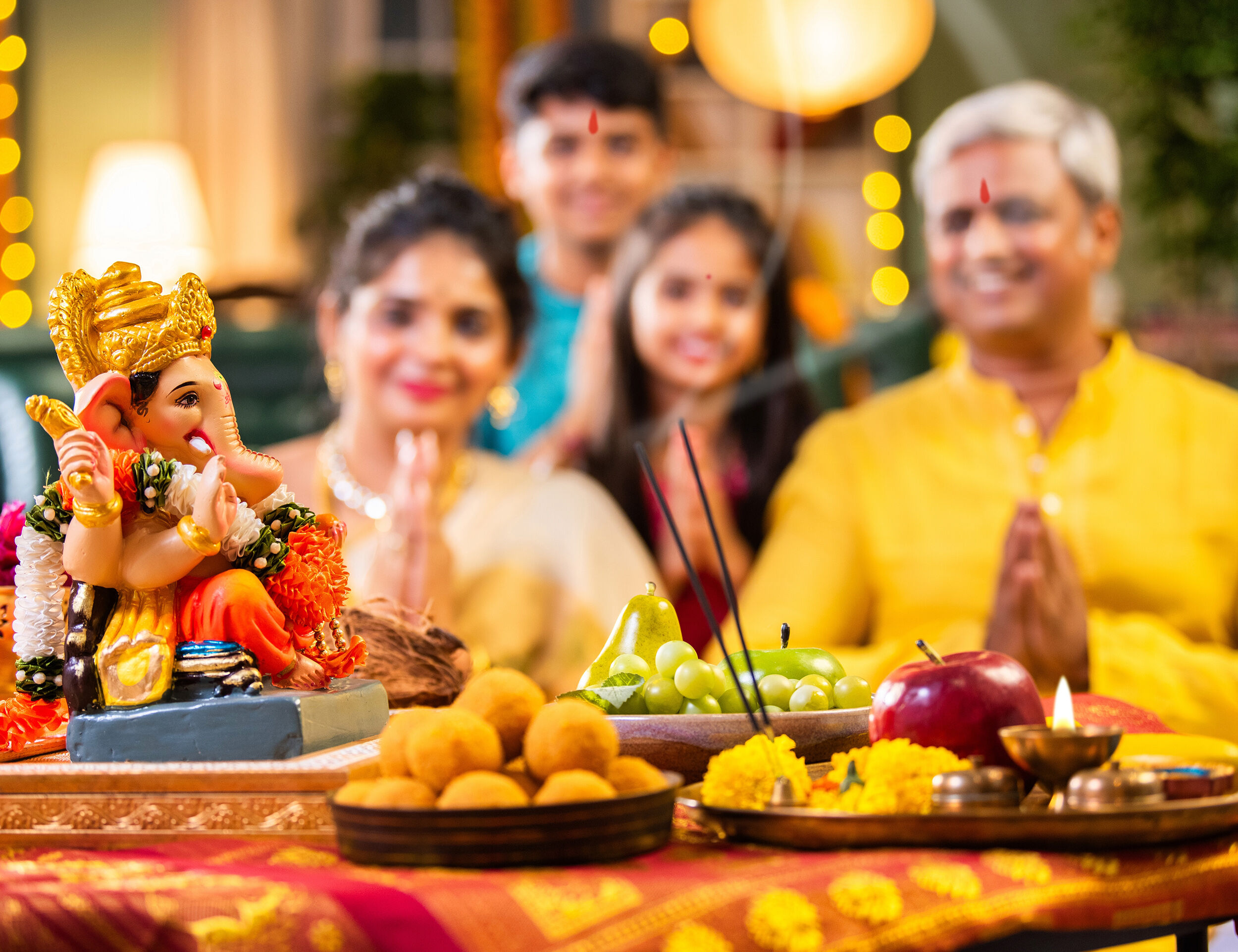 Happy Indian family celebrating Ganesh Chaturthi at home with a decorated Lord Ganesha idol, traditional sweets, fruits, flowers, and incense offerings.
